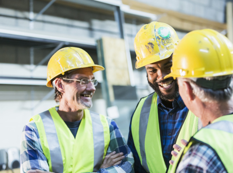 Three construction workers wearing yellow hard hats and high vis vests, standing on a building site, smiling and talking