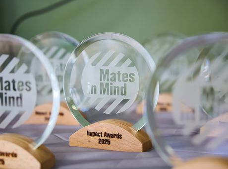 Circular glass trophies with the Mates in Mind logo engraved on it, and standing on a wooden block with Impact Awards 2025.
