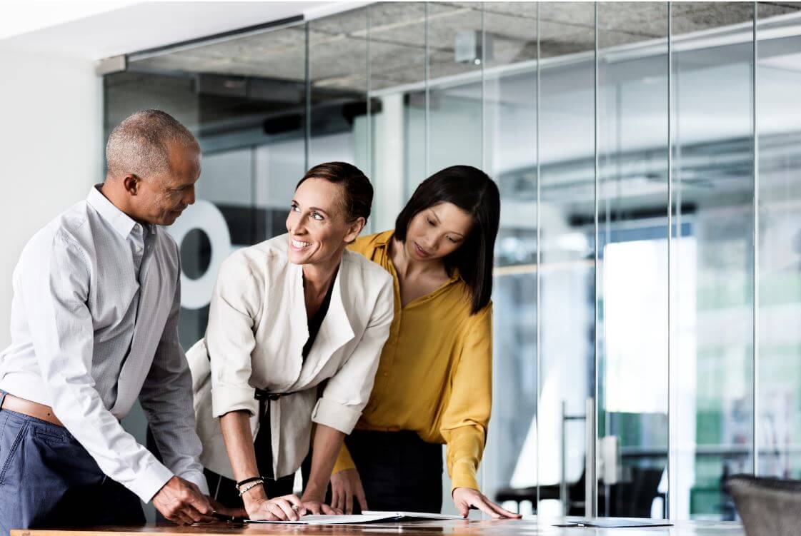 Three employees standing in an office with a glass wall, discussing a Mates in Mind report which is open on the desk in front of them